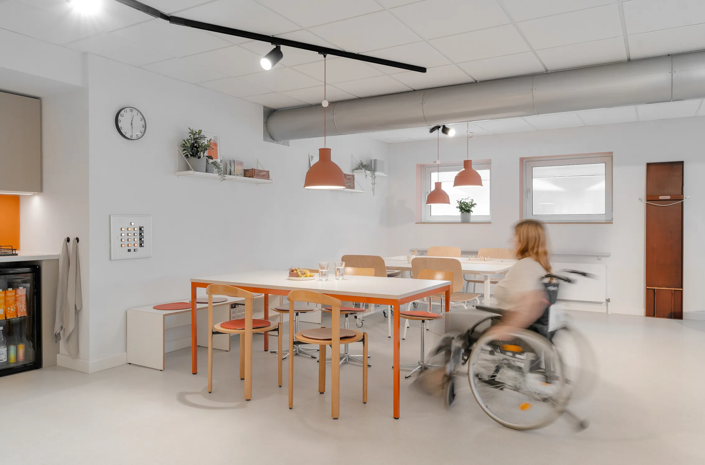 Ein Raumkonzept von Cajodesing, Rollstuhlgeeignet. Ein Tisch mit mehreren Stühlen, eine Person im Rollstuhl bewegt sich an den Tisch.