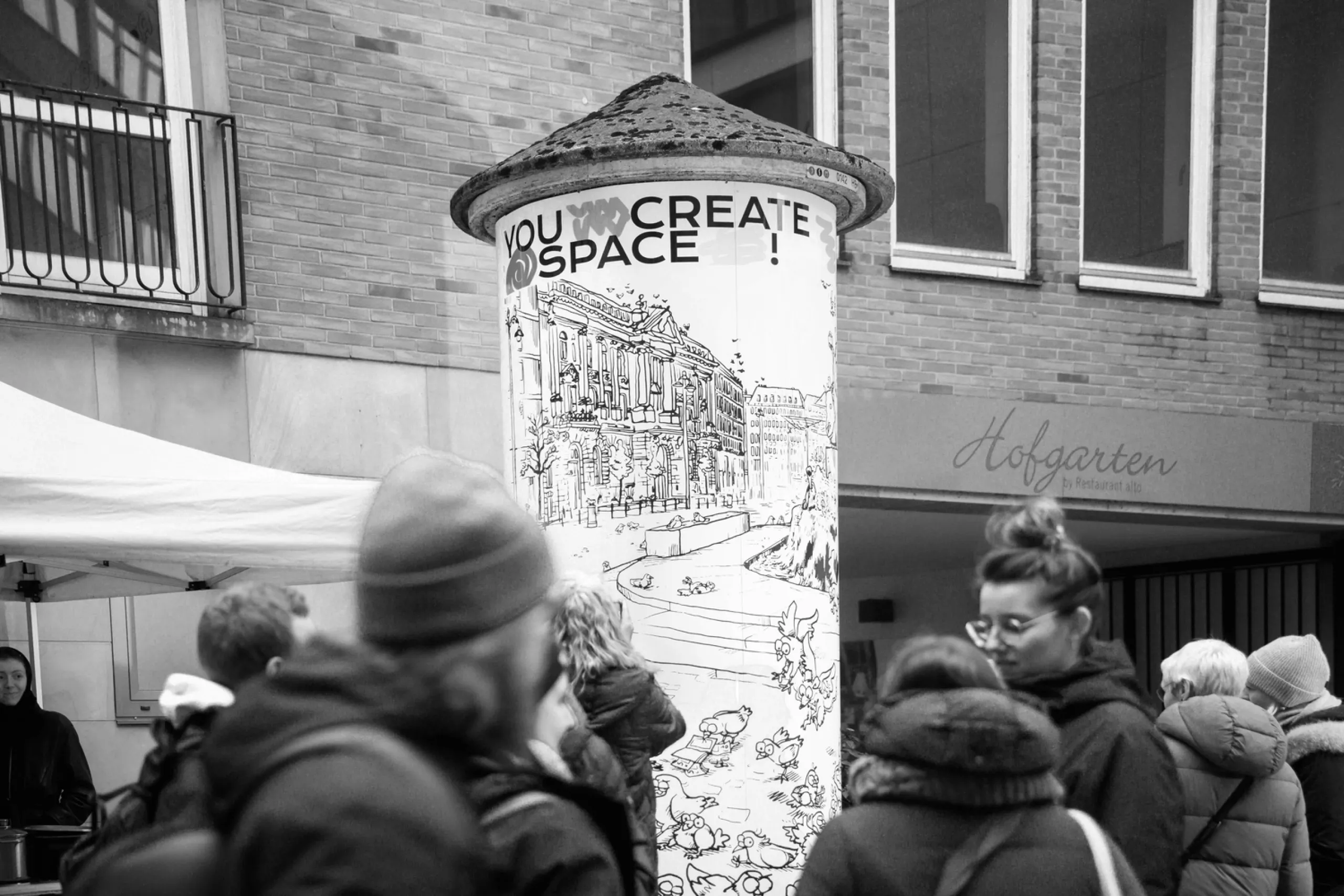 Schwarz-weiß-Fotografie einer Litfaßsäule in der Stadt, beklebt mit einer Illustration voller Tauben. Menschen mit Winterkleidung stehen davor, darüber steht der Schriftzug ‚You create space‘.