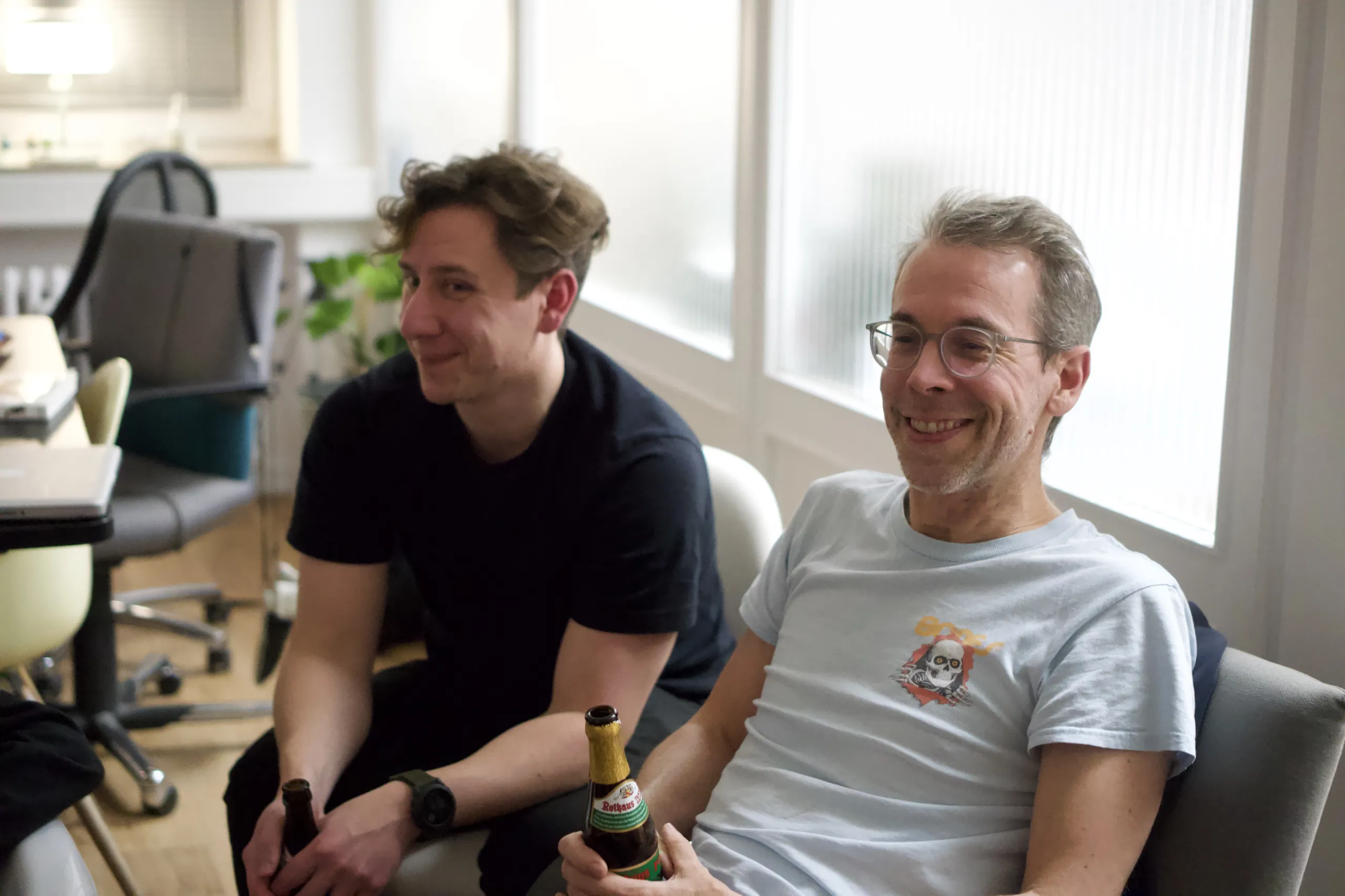 Zwei lächelnde Männer im Atelier Hund, sie sitzen, trinken Bier und schauen auf etwas, das nicht im Bild ist.