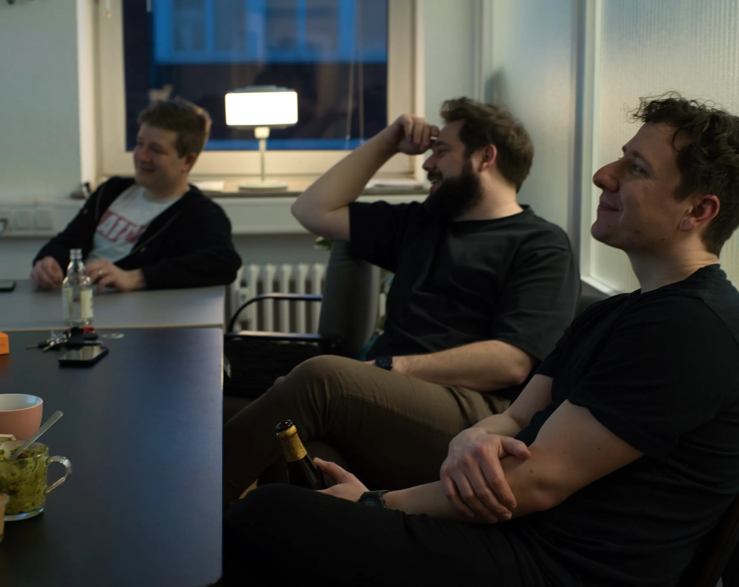 Drei lächelnde Männer im Atelier Hund, sie sitzen, trinken Bier und schauen auf etwas, das nicht im Bild ist.